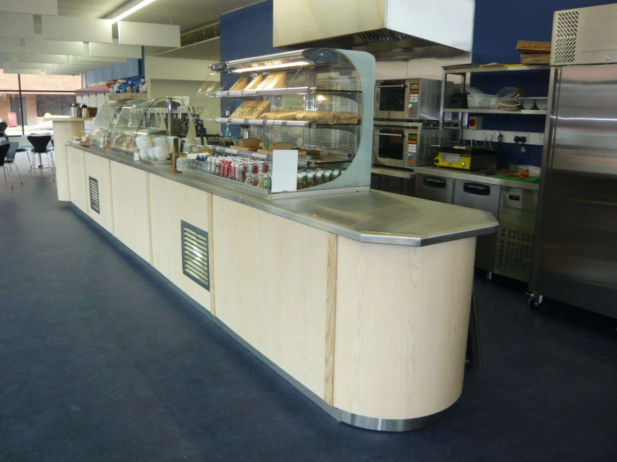 A long, curved wooden school or office cafeteria servery counter with a stainless steel worktop, glass sandwich display unit, and commercial kitchen ovens in the background.
