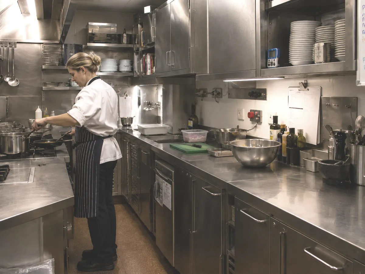 A female chef in a white uniform and striped apron cooking at a commercial hob next to long stainless steel preparation worktops.