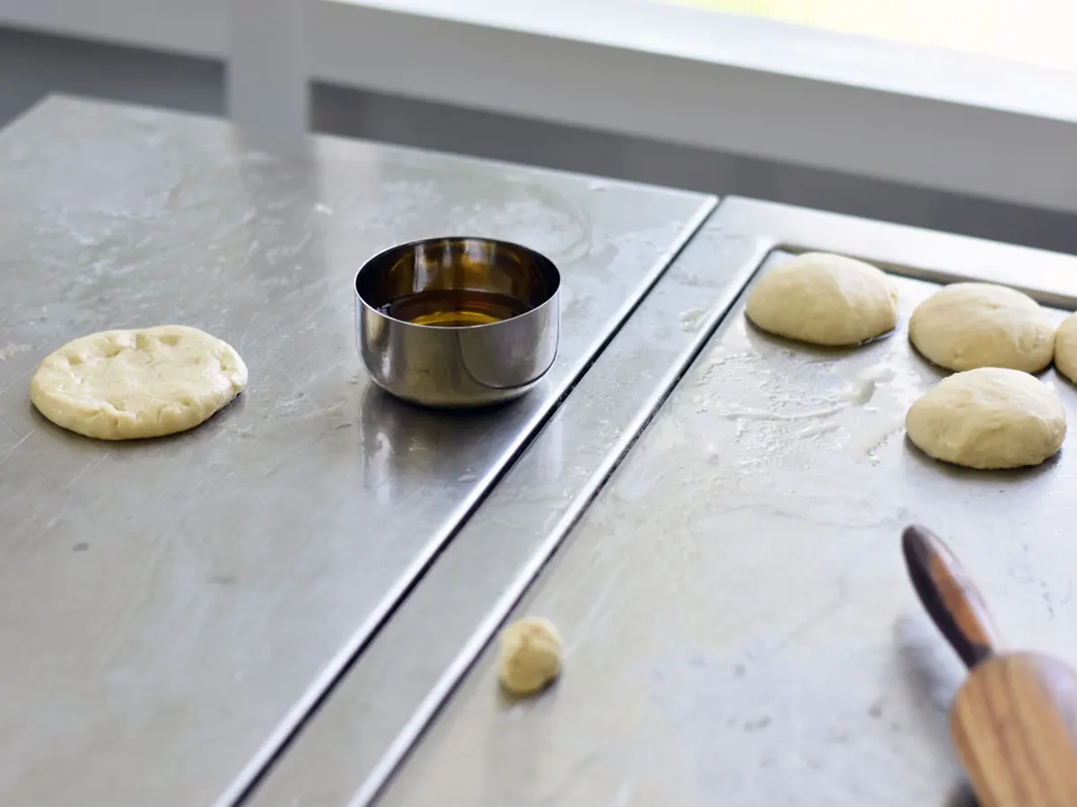 Portions of uncooked bread dough being shaped and prepared on a smooth stainless steel worktop alongside a small metal bowl of oil and a wooden rolling pin.