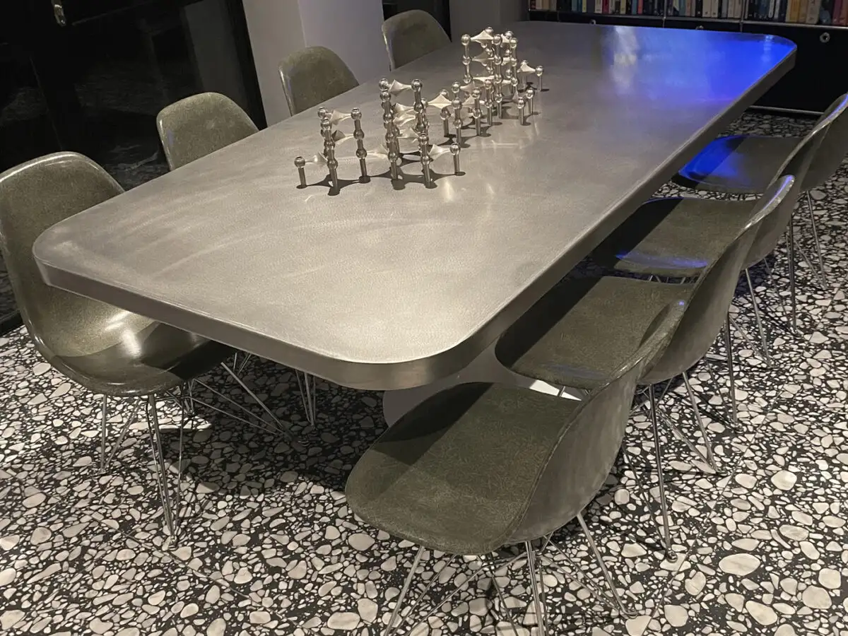 Large rectangular stainless steel dining table with rounded corners, surrounded by dark green modern chairs on a black and white terrazzo tile floor.